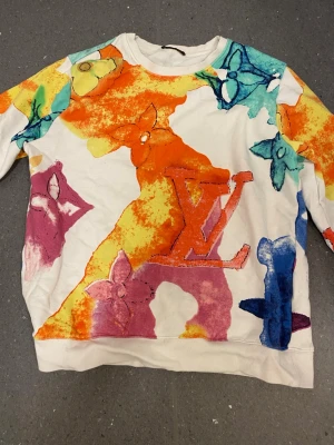 Färgglad Louis Vuitton sweatshirt - Unik sweatshirt från Louis Vuitton med färgstarkt mönster i orange, rosa, blått, gult och grönt. Tröjan har LV-logga och blommor tryckta över hela plagget. Rund halsringning och långa ärmar. Perfekt statement piece för dig som gillar streetwear och lyx.