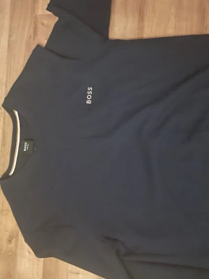 Mörkblå tröja från BOSS - Snygg mörkblå tröja från BOSS med diskret logga broderad på bröstet. Tröjan har rund halsringning med en stilren rand i vitt och beige. Perfekt för en clean och klassisk look.