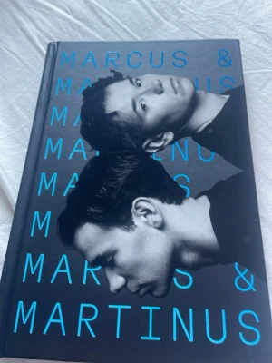 Marcus & Martinus - Två röster, en historia - En svart hårdbunden bok med blå text och foto på Marcus & Martinus på omslaget. Boken handlar om tvillingarnas resa till att bli popstjärnor och deras personliga berättelser om livet i rampljuset. Perfekt för dig som gillar biografier och musik.