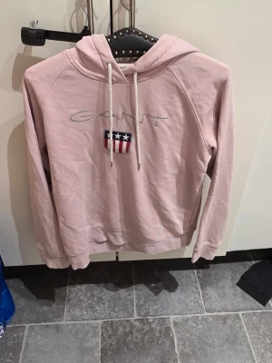 Ljusrosa hoodie från Gant - Säljer en ljusrosa hoodie från Gant med broderad logga och amerikanskinspirerat märke på bröstet. Hoodien har vit dragsko i huvan och mjukt material, perfekt för en avslappnad stil. Klassisk passform och ribbade muddar.