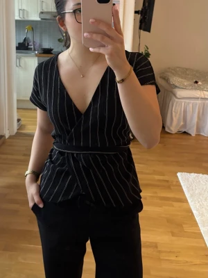 Gina Tricot omlottopp, XS - Säljer en omlottopp från Gina Tricot. Den är i bra skick med inga tecken på slitage. Den är lätt och materialet är av 90% polyester. Det tillkommer ett snöre man kan knyter runt midjan. Finns inte att köpa längre och har svart färg med ljusa randiga detaljer. 