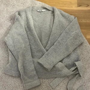 Grå omlottkofta från NA-KD - Mysig grå stickad omlottkofta från NA-KD i storlek S. Koftan har långa ärmar, v-ringning och knytband i midjan för en snygg passform. Perfekt att slänga på sig när det är lite kyligt ute. Köptes för 400kr och säljs för 299 för har bara används 1 gång ( pris kan diskuteras)