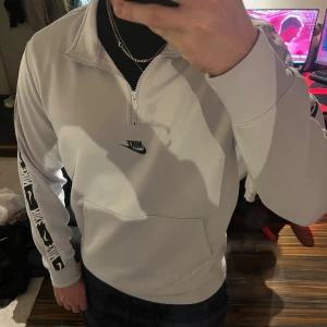 Vit Nike half zip hoodie - Säljer en fräsch vit hoodie från Nike med half zip och svart Nike-logga på bröstet. Ärmen har coolt svartvitt mönster och tröjan har en stor magficka. Perfekt för dig som gillar sportig och clean stil. Funkar även väldigt bra som träningströja!