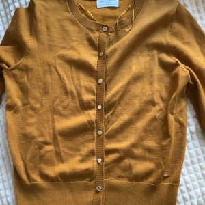 Gul kofta från Holly & White - Gul kofta från Holly & White med rund halsringning och knappar framtill. Långärmad modell i mjuk bomull, perfekt att slänga över en t-shirt eller klänning. Ribbstickade muddar vid ärmslut och nederkant för en snygg passform. Pris går att diskutera.