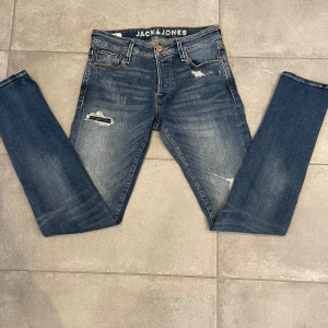 Jack and Jones jeans - Snygga blå jeans från Jack & Jones med slitna detaljer och en modern look. De har en knappgylf och klassisk femficksdesign. Perfekta för en avslappnad stil. Storlek W30 L34
