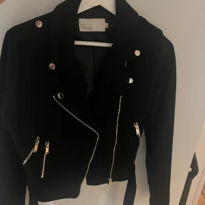 Svart bikerjacka från NLY Trend - Svart mocka bikerjacka från NLY Trend med silvriga dragkedjor, knappar och bälte i midjan. 