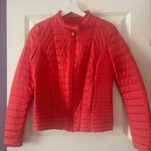 Röd pufferjacka från Guess - Snygg röd pufferjacka från Guess med quiltad design och dragkedja framtill. Jackan har en smal passform, hög krage och diskret logga bak. Perfekt för dig som vill sticka ut med färg i höst och vinter.