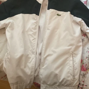 Vit och svart vindjacka från Lacoste - Snygg vindjacka från Lacoste i vit med svart axelparti och klassisk krokodillogo på bröstet. Jackan har dragkedja framtill, två sidofickor och elastisk nederkant. Perfekt för dig som gillar sportig och clean stil.