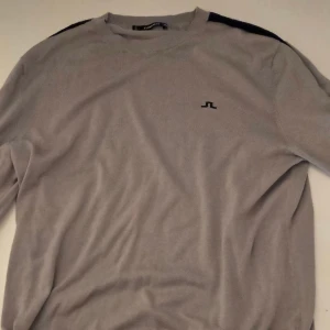 Grå tröja med marinblå ränder J.Lindeberg - Snygg grå tröja från J.Lindeberg med marinblåa ränder längs ärmarna och logga på bröstet. Tröjan har rund halsringning och är långärmad.