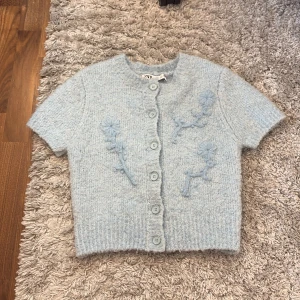 Ljusblå stickad kofta från Zara - Superfin ljusblå stickad kofta från Zara med kort ärm och broderade blommor framtill och baktill. Koftan har rund hals, knappar hela vägen och ribbad nederkant. 