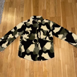 Camo teddyjacka i fleece - Säljer en oversized teddyjacka med camomönster i svart, beige och grönt. Jackan är gjord i mjuk fleece, har krage, bröstficka och knäppning framtill. Perfekt för dig som vill sticka ut med en cool och mysig look. Storlek 134-140