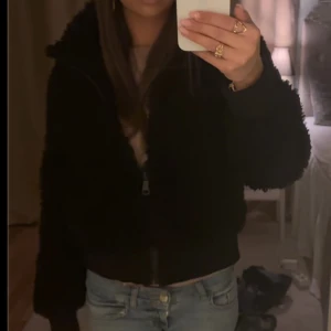 Svart teddyjacka med dragkedja - Mysig svart teddyjacka med fluffig känsla och dragkedja framtill. Jackan har en lite croppad passform och långa ärmar, perfekt för att hålla värmen och ändå se cool ut. Enkel att matcha med jeans eller kjol för en chill look.