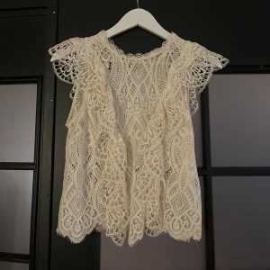 Vit spetsblus från Zara - Superfin vit blus från Zara i storlek XS, med detaljerad spets och volang vid axlarna. Blusen är ärmlös och har ett romantiskt mönster med snygga broderier. Perfekt för dig som gillar en söt och trendig look.