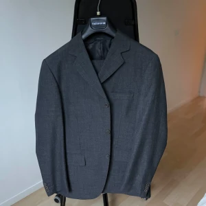 Mörkgrå kostym – Medi, Made in Italy (stl 48) - Elegant mörkgrå kostym från det italienska kvalitetsmärket Medi. En tidlös helkostym i klassisk design som passar lika bra på kontoret som vid fest och högtidliga tillfällen.  Storlek: 48  Färg: Mörkgrå  Modell: F013A  Tillverkad i Italien  Skick: Helt ny med tags kvar