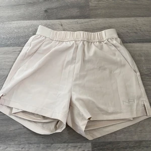 Beige träningsshorts från New Apparel - Snygga beige träningsshorts från New Apparel med elastisk midja och diskret logga på benet. Shortsen har innerbyxa, slits i sidorna och är tillverkade i mjuk polyester med skön passform. Perfekta för gymmet eller löprundan.