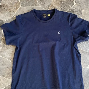Marinblå t-shirt från Polo Ralph Lauren - Snygg marinblå t-shirt från Polo Ralph Lauren med klassisk rund hals och korta ärmar. T-shirten är i mjuk bomull och har den ikoniska vita Polo-loggan broderad på bröstet. Perfekt basic med stilren look och skön passform.