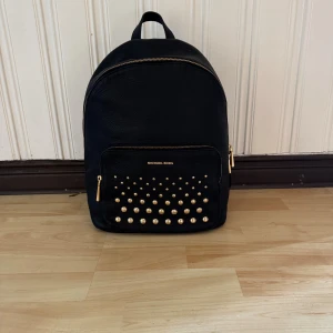 Svart ryggsäck Michael Kors med nitar - Snygg svart ryggsäck från Michael Kors i skinn med guldfärgade nitar på frontfickan och guldfärgade dragkedjor. Rymligt huvudfack, innerficka med dragkedja och justerbara axelremmar. Den har vara stått så den har prislappen kvar. 