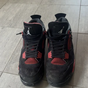 Nike Air Jordan 4 svart/röd sneakers - Säljer ett par Nike Air Jordan 4 sneakers i svart och rött med klassisk Jumpman-logga på plösen och hälen. Skorna har svarta snören, meshdetaljer och röda accenter. Ovandelen är i mocka och syntet, med en robust sula och cool retrostil.