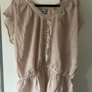 Beige siden blus med paljetter Noa Noa - Supersöt beige blus från Noa Noa med glittriga paljetter längs halsringningen och framtill. Blusen har korta ärmar, resår i midjan och är tillverkad i ett tunt, lätt material som känns luftigt och skönt. Materialet är siden