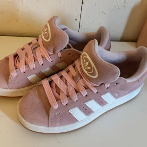 Adidas Campus rosa sneakers mocka - Snygga Adidas Campus sneakers i ljusrosa mocka med vita klassiska ränder och rosa skosnören. Skorna har en vit sula och rund tå, samt Adidas logga på plösen. Perfekta för dig som vill ha en fräsch och trendig look.