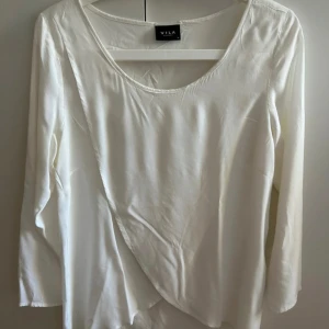 Vit omlottblus från Vila - Säljer en stilren vit omlottblus från Vila med lång ärm och rund halsringning. Blusen har en snygg omlottdetalj framtill som ger en cool och modern look. Materialet är mjukt och har ett lätt fall, perfekt för att lyfta din outfit