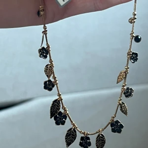 Guldigt halsband med blommor från Pilgrim - Supersnyggt halsband från Pilgrim i guldfärgad metall med svarta blom- och lövberlocker. Kedjan är tunn och dekorerad med små detaljer som ger en cool och unik vibe. Perfekt för dig som gillar att sticka ut med accessoarer med blommigt tema.