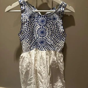 Söt känning  - Superfin vit kortklänning med blå broderade detaljer och mönster på överdelen. Klänningen har ärmlös design och rund hals, med en lätt och luftig kjol. Perfekt för varma dagar och har en unik, ungdomlig vibe med de coola broderierna.
