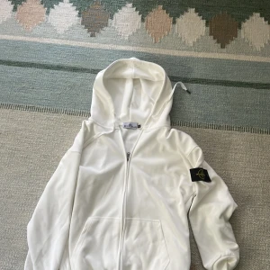 Vit hoodie från Stone Island - Säljer en clean vit hoodie från Stone Island med dragkedja och klassisk logga på ärmen. Tröjan har huva, fickor framtill och är tillverkad i mjukt bomullsmaterial. Perfekt för dig som gillar streetwear och vill ha en stilren look. Tröjan är oanvänd.