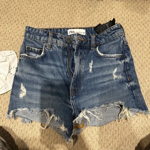  jeansshorts från Zara - Säljer ett par blå jeansshorts från Zara med fransig och rå kant nertill. Shortsen har klassisk femficksdesign, slitna detaljer och knapp- samt dragkedja framtill. Perfekt för dig som gillar en avslappnad och cool stil.