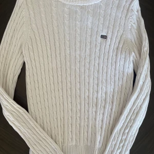 Vit kabelstickad tröja från Lexington - Vit kabelstickad tröja från Lexington med rund halsringning och diskret logga på bröstet. Tröjan har lång ärm och är gjord i mjuk bomull, perfekt för lager-på-lager och en clean look.