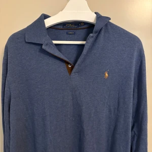 Blå långärmad pikétröja Polo Ralph Lauren - Helt oanvänd  blå långärmad pikétröja från Polo Ralph Lauren i bomull. Tröjan har klassisk krage, knappslå med brun detalj och den ikoniska broderade loggan på bröstet. Classic fit för en avslappnad stil.
