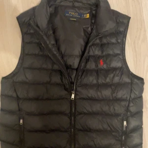 Svart dunväst från Polo Ralph Lauren - Snygg svart dunväst från Polo Ralph Lauren med klassisk quiltad design och röd logga på bröstet. Västen har dragkedja framtill och två fickor med dragkedja. Perfekt att slänga över en hoodie eller tröja för en sportig look. Nypriset på västen ligger runt 2800, säljer för lägre pris pågrund av ett hål i baksidan av västen. Det är ett hyfsat litet hål men det kan ändå märkas av. Går säkert att fixa dock! 