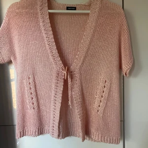 Ljusrosa stickad kofta från Gerry Weber - Supersöt ljusrosa stickad kofta från Gerry Weber med korta ärmar och djup v-ringning. Koftan har knytning framtill och två dekorativa fickor. Perfekt för lager-på-lager och ger en chill vibe till din outfit.