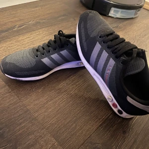 Svarta Adidas sneakers med mesh - Snygga svarta Adidas sneakers med klassiska tre ränder i grått på sidorna. Ovandelen är i ventilerande mesh och skorna har snörning samt platt vit sula. Perfekta för en sportig och avslappnad stil.