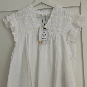 Vit topp/blus från Gina  - Superfin vit blus från Gina med volangärmar och spetsdetaljer. Lätt och luftig bomull, perfekt för varma dagar. Söt v-ringning och romantisk känsla, passar dig som gillar en enkel men söt stil.
