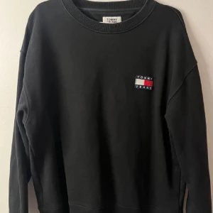 Svart sweatshirt från Tommy Jeans - Svart sweatshirt från Tommy Jeans med klassisk logga broderad på bröstet i vitt, rött och blått. Tröjan har rund halsringning och långa ärmar. Perfekt för en chill och stilren look.