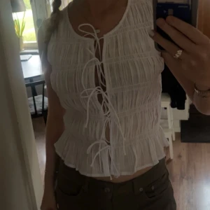 Vit blus med snörning framtill - Supersöt vit blus med flera snörningar framtill och lätt rynkad struktur. Modellen är ärmlös och har en croppad passform med volangkant nedtill. Perfekt för varma dagar och ger en romantisk vibe.