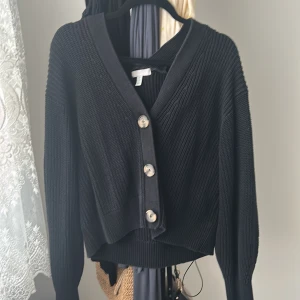 Svart stickad kofta från H&M - Svart stickad kofta från H&M i storlek S. Koftan har grov ribbstickning, v-ringning och stora, snygga knappar framtill. Perfekt att slänga över en t-shirt eller klänning för en chill look.