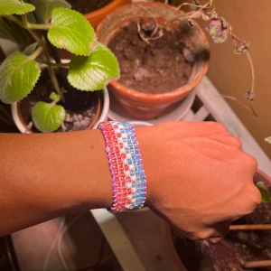 Färgglatt pärlarmband med snörning - Unikt handgjort armband med pärlor i rosa, blått och vitt, trädda på lila snöre. Armbandet har en bred design och justerbar knytning för perfekt passform. Perfekt för dig som vill ha en färgklick på handleden och gillar personliga accessoarer.