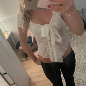 Vit blus med knyt och spetsdetaljer - Supersöt vit blus med korta ärmar och stor knytning framtill. Tunt, genomskinligt tyg med små broderade blommor och vågig nederkant. Perfekt för dig som gillar romantisk och feminin stil. Säljs i storlek S.