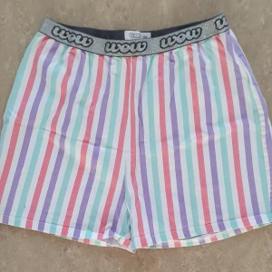 Shorts - Färgglada shorts från Wow med vertikala ränder i lila, rosa, turkos, röd och vit. De har en bred, glittrig silverfärgad resår med Wow-logga runt midjan. Modellen är loose fit och midwaist, perfekta för chill dagar. Materialet känns mjukt och lätt, troligen bomull eller polyester.