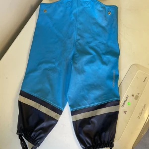 Blå galonbyxor från Didriksons stl 80 - Blå galonbyxor från Didriksons med svarta partier och reflexband nedtill. Byxorna har resår i bensluten och justerbara fothällor. Materialet är 100% polyester med polyuretanbeläggning, perfekta för regniga dagar.