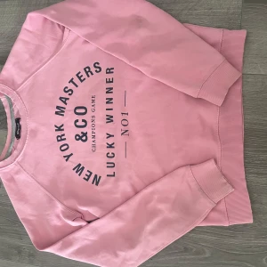 Rosa sweatshirt New York Masters - Rosa sweatshirt med svart tryck framtill där det står 'New York Masters & Co Lucky Winner No1'. Tröjan har rund hals, långa ärmar och ribbade muddar vid ärmslut och nederkant. Perfekt för en chill och avslappnad stil.