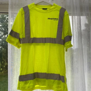 Gul varsel tröja - Neongul t-shirt med grå reflexband från Praktiska Gymnasiet. Klassisk rund hals och korta ärmar, perfekt för dig som vill synas. Tryck med skolans logga på bröstet. Materialet känns lätt och bekvämt, passar dig som gillar funktionella plagg.