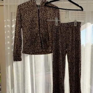 Leopardmönstrade byxor med ziphoodie från Gina - Säljer ett par leopardmönstrade byxor med en ziphoodie från Gina Tricot i mjukt, stretchigt material. De är beige med bruna och svarta fläckar, har normal passform och resår i midjan. Perfekta för dig som vill sticka ut med en cool look.
