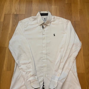 Vit skjorta från Polo Ralph Lauren - Klassisk vit skjorta från Polo Ralph Lauren med broderad logga på bröstet. Skjortan har lång ärm, knappar hela vägen och button down-krage. Tillverkad i bomull och har en stilren, tidlös look som funkar till många olika outfits. Den är krympt från en L till en M i storlek. Inga skador eller annat.