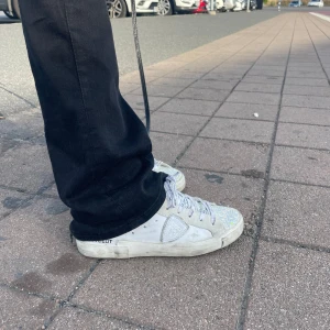 Glittriga sneakers från Philippe Model - Säljer ett par vita sneakers från Philippe Model med glittriga detaljer på tån och silvriga snören med logga. Skorna har en klassisk låg profil, rund tå och platt sula. Märkets sköldlogga syns på sidan och sulan är i gummi. Perfekta för dig som vill sticka ut lite extra.