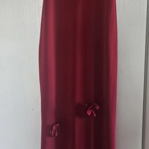Vinröd långklänning med rosetter - Säljer en elegant vinröd långklänning med dekorativa rosetter nedtill. Klänningen har en rak och stilren siluett, perfekt för dig som vill sticka ut med en enkel men snygg look. Materialet känns mjukt och har ett fint fall. Jag som haft den är 1, 62 och jag hade en klack på ca 6 cm när jag använde klänningen. Klänningen är endast använd en gång. Nypris är 1400:-. 