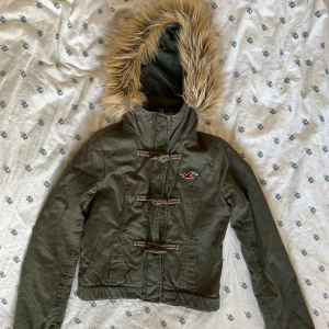 Vintage cropped Olivgrön jacka med fuskpäls från Hollister - Snygg vintage cropped olivgrön jacka från Hollister med huva och fuskpäls. Jackan har tre träknappar framtill, broderad logga på bröstet och är fodrad med mjuk teddy. Yttermaterial i bomull och foder i polyester. Perfekt för kyliga dagar! Några små hål på höger armen. 