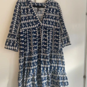 Blåvit mönstrad klänning Vero Moda - Supersnygg blå och vit klänning från Vero Moda med grafiskt mönster, v-ringning och trekvartsärmar. Lätt och luftig modell med avslappnad passform, perfekt för varma dagar. Klänningen har en bohemisk vibe och är tillverkad i mjukt material.